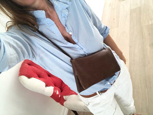 Woman standing wearing an envelope shaped crossbody bag in distressed brown leather. The bag is made of two separate “envelopes” its size being close to a standard #10