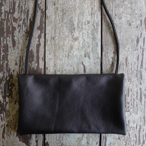Envelope shaped black leather crossbody bag lays on a table top. The size of this bag is close to that of a standard #10 envelope.