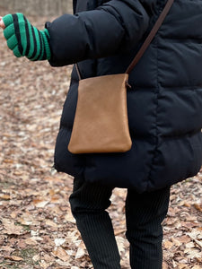 woman stands wearing x-small tan leather crossbody bag. its bottom corners are rounded.