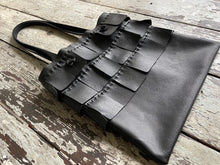 Load image into Gallery viewer, A black leather bag with three rows of wide black leather hand sewn fringe covering 1/3 of the small-size bag starting at the top lays flat on a table top its shoulder.
