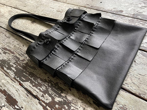 A black leather bag with three rows of wide black leather hand sewn fringe covering 1/3 of the small-size bag starting at the top lays flat on a table top its shoulder.