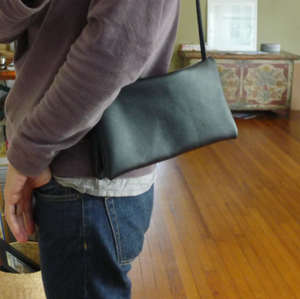 Person standing wearing an envelope shaped black leather crossbody bag. The bag is made of two separate “envelopes” its size being close to a standard #10.