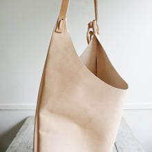 Load image into Gallery viewer, Side view of a shoulder length bag made in natural vegetable tanned leather. The top of the bag dips down into a half-circle shape. Leather straps are tied and knotted at the top left and right sides. The bottom half of the bag is a rectangular shape and the body is flat.
