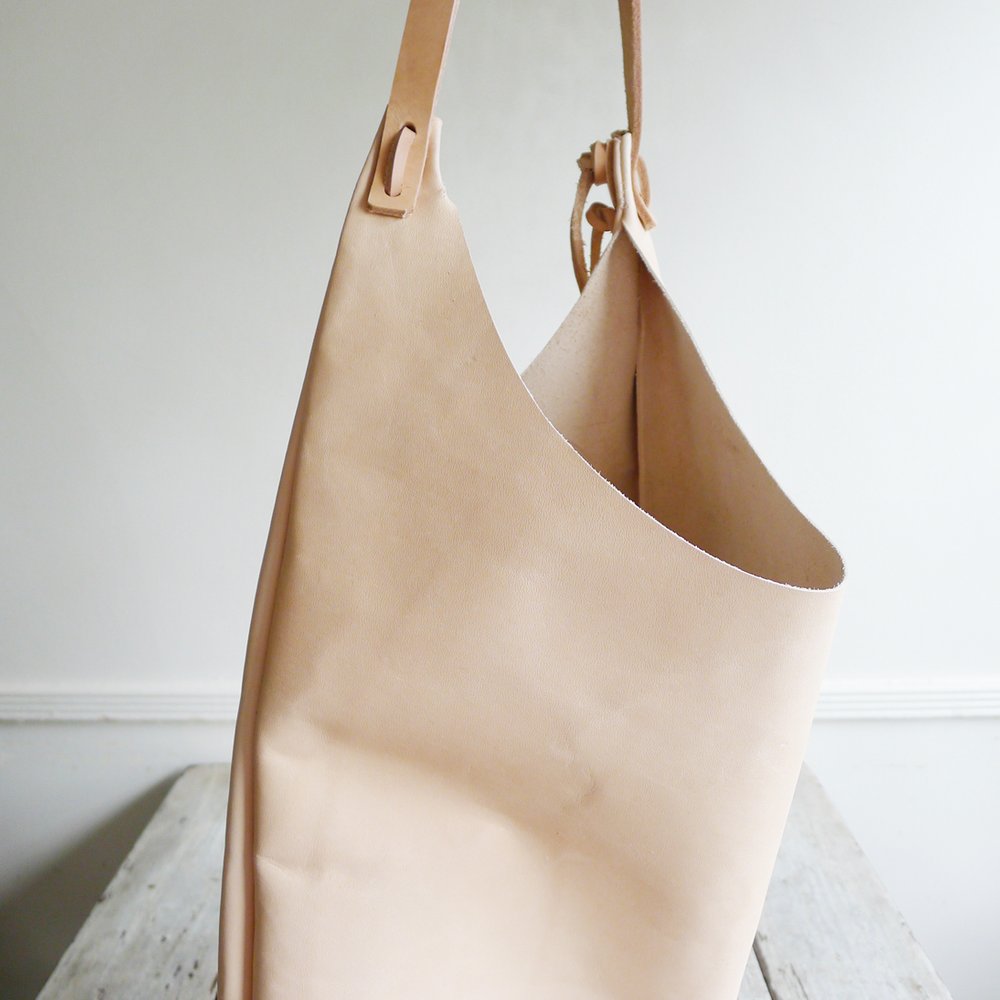 Side view of a shoulder length bag made in natural vegetable tanned leather. The top of the bag dips down into a half-circle shape. Leather straps are tied and knotted at the top left and right sides. The bottom half of the bag is a rectangular shape and the body is flat.