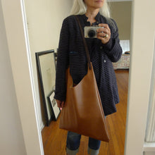 Load image into Gallery viewer, Woman is wearing a crossbody length leather bag. The top of the bag dips down into a half-circle shape. Leather straps are tied and knotted at the top left and right sides. The bottom half of the bag is a rectangular shape and the body is flat. The leather looks supple. This bag can be ordered in black, distressed brown and natural veg tan leather and is no longer available in the pictured tan leather. 
