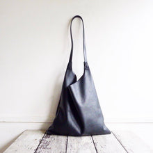 Load image into Gallery viewer, An all-black leather bag sits bottom up on a table. both the left and the right sides at the top of the bag have a 3/4” area where each end of the leather strap is tied and knotted. The body/bottom half of the bag is a rectangular shape. The leather looks supple.  The leather looks supple. this bag is artisan made and handcrafted.  The leather looks supple. this bag is artisan made and handcrafted.
