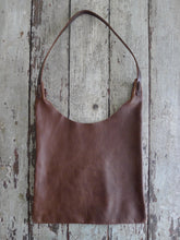 Load image into Gallery viewer, An all-brown shoulder length leather bag lays flat on a table top. The top of the bag dips down into a half-circle shape. Leather straps are tied and knotted at the top left and right sides. The bottom half of the bag is a rectangular shape and the leather body looks supple
