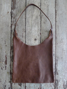An all-brown shoulder length leather bag lays flat on a table top. The top of the bag dips down into a half-circle shape. Leather straps are tied and knotted at the top left and right sides. The bottom half of the bag is a rectangular shape and the leather body looks supple