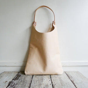 Front view of a shoulder length bag made in natural vegetable tanned leather. The top of the bag dips down into a half-circle shape. Leather straps are tied and knotted at the top left and right sides. The bottom half of the bag is a rectangular shape and the body is flat.