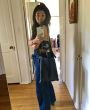 Load image into Gallery viewer, A dark haired female stands wearing a narrow and flat rectangular shaped black leather bag over her shoulder. The matching leather strap is tied and knotted to each side of the bag. The bag has a large front pocket. The leather looks supple.
