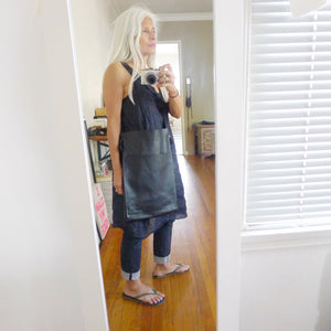 A white haired female stands wearing a narrow and flat rectangular shaped black leather bag over her shoulder. The matching leather strap is tied and knotted to each side of the bag. The bag has a large front pocket. The leather looks supple.