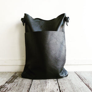 a narrow and flat rectangular shaped black leather bag lays on a table top. The leather looks supple. The black leather strap is tied and knotted to each side of the bag. The front of the bag has a large outside pocket