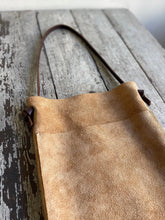 Load image into Gallery viewer, a narrow and flat rectangular shaped tan suede leather bag lays on a table top. The suede looks supple. The dark brown leather strap is tied and knotted to each side of the bag. The front of the bag has a large outside pocket. 
