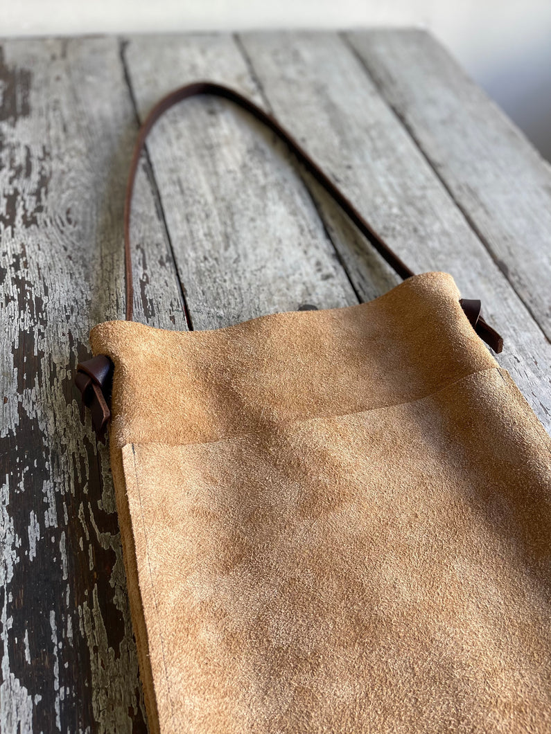 a narrow and flat rectangular shaped tan suede leather bag lays on a table top. The suede looks supple. The dark brown leather strap is tied and knotted to each side of the bag. The front of the bag has a large outside pocket. 