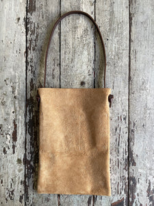 a narrow and flat rectangular shaped tan suede leather bag lays on a table top. The suede looks supple. The dark brown leather strap is tied and knotted to each side of the bag. The front of the bag has a large outside pocket. 
