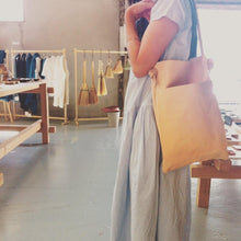 Load image into Gallery viewer, A woman standing in a store wears a narrow and flat rectangular shaped natural veg tan leather bag over her shoulder. The leather patina is a soft peachy blush color. The matching leather strap is tied and knotted to each side of the bag. The front of the bag has a large outside pocket.  
