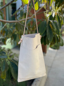 a narrow and flat rectangular shaped natural veg tan leather bag hangs from a tree branch. The leather patina is a soft peachy blush color. The matching leather strap is tied and knotted to each side of the bag. The front of the bag has a large outside pocket.