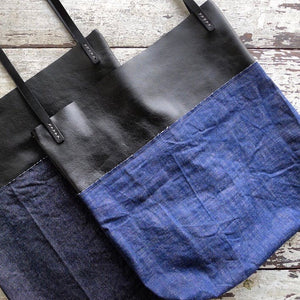 laying on a table top are two bags made from a combination of blue denim & black leather material. Two shoulder length black leather straps are hand sewn to the top of the bag using grey linen yarn and are attached on each side. This bag is artisan made and handcrafted.
