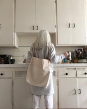 Load image into Gallery viewer, Back to the viewer, a white haired woman in kitchen wears a combination of natural denim &amp; natural-veg tan leather bag on her back (like you would wear a backpack). This bag is artisan made and handcrafted.
