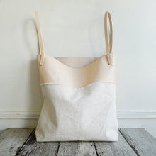 Load image into Gallery viewer, sitting on a table top is a bag made using a combination of natural denim &amp; natural-veg tan leather. Two shoulder length veg tan leather straps are hand sewn using white linen yarn to the top of the bag attached on each side. This bag is artisan made and handcrafted.
