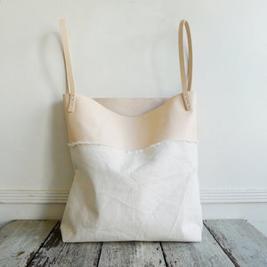sitting on a table top is a bag made using a combination of natural denim & natural-veg tan leather. Two shoulder length veg tan leather straps are hand sewn using white linen yarn to the top of the bag attached on each side. This bag is artisan made and handcrafted.