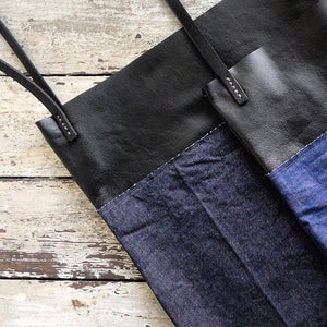 a close up two bags made from a combination of blue denim & black leather material. Two shoulder length black leather straps are hand sewn to the top of the bag using grey linen yarn and are attached on each side. This bag is artisan made and handcrafted.