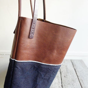 Side view of a combination of dark blue denim & distressed brown leather bag. Two shoulder length brown leather straps are hand sewn to the top of the bag using grey linen yarn and are attached on each side.This bag is artisan made and handcrafted.