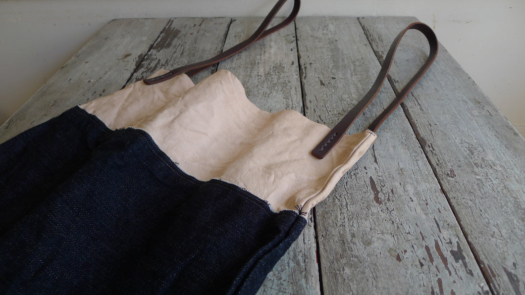 sitting on a table top is a bag made using a combination of dark denim & natural-veg tan leather. Two shoulder length brown leather straps are hand sewn using grey linen yarn to the top of the bag attached on each side. The bag has been washed. This bag is artisan made and handcrafted.