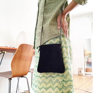 woman stands wearing x-small black suede bag its bottom corners are rounded. strap length can be worn crossbody. This bag is artisan made and handcrafted.