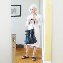 Load image into Gallery viewer, Woman standing wearing a crossbody black leather bag. Drawstring style— the black leather ties cinch in for closure. Ties are removable to wear bag to its fully extended width. Made with knotted and adjustable straps. The bags leather looks soft and supple.
