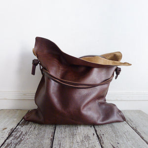 Close up of distressed brown leather bag. Drawstring style— the brown leather ties cinch in for closure. Ties are removable to wear bag to its fully extended width. Made with knotted and adjustable strap. The top of the bag is folded open exposing its suede interior. The bags leather looks soft and supple.
