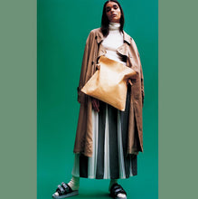 Load image into Gallery viewer, Woman standing wearing a crossbody natural vegetable tanned leather bag (with a light patina). Drawstring style— the veg tan leather ties were removed to wear the bag to its fully extended width. Made with knotted and adjustable straps.

