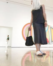 Load image into Gallery viewer, A woman standing (in an art gallery surrounded by art) holding a small black suede bag fully gathered at the top tied with a black leather tie knotted for closure. It’s short black strap is shoulder length. The bag has a center seam and a gusset bottom.
